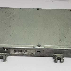 Pode incluir: Uma unidade de controlo eletrónico (ECU) prateada com formato retangular. A ECU tem uma caixa metálica com parafusos e suportes de montagem visíveis. Um código de barras e texto são visíveis numa etiqueta.