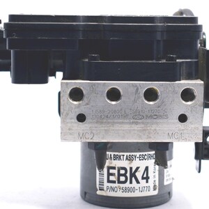 Op de afbeelding: Zwart en zilver auto-onderdeel met meerdere componenten, waaronder een beugel, een cilindrisch lichaam en een rechthoekige metalen plaat met ronde gaten. Het onderdeel heeft de tekst "EBK4" en "P/NO 58900-1J770".