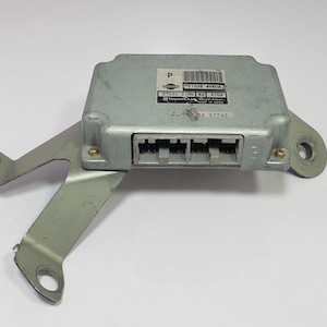 May include: A silver electronic control unit (ECU) with a metal bracket. The ECU is rectangular with connectors and a label with text and a barcode. The bracket has holes for mounting. The unit is likely for a vehicle.