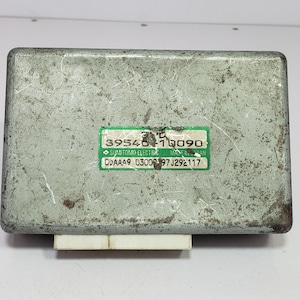 Op de afbeelding: Een rechthoekig, verweerd, grijs metalen elektronisch component met een witte connector. Een groene en witte sticker toont de nummers "39546-10090" en andere tekst. Het component is waarschijnlijk voor automotive of industrieel gebruik.