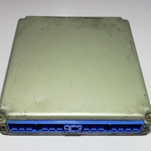 以下が含まれることがあります： 青いコネクタが付いた、長方形の銀色の電子制御ユニット（ECU）。ECUは、わずかに摩耗した外観で、目に見える傷があります。青いコネクタは片側にあり、2本のネジで固定されています。