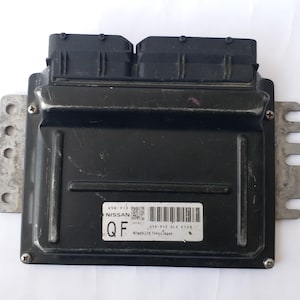 May include: A black Nissan engine control unit (ECU) with two black connectors. The rectangular unit has a label with the Nissan logo and other text. Metal mounting brackets are attached to the sides. Made in Tokyo, Japan.