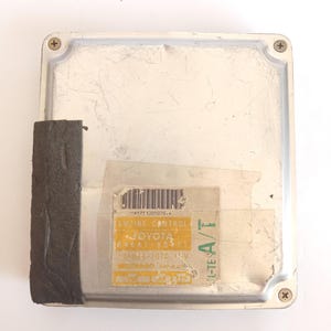 May include: A silver engine control unit with a black foam pad attached to the side. A yellow and white sticker with text and a barcode is affixed to the front. The sticker reads "ENGINE CONTROL TOYOTA".