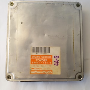 May include: A rectangular, silver-colored engine control unit with a label that reads "ENGINE CONTROL TOYOTA." The label also includes the text "DENSO MADE IN JAPAN" and other numerical data. The unit is secured with four screws.