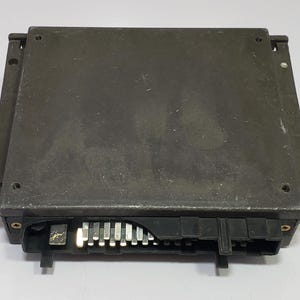 May include: A rectangular, dark gray electronic component with a series of metal pins and black connectors. The component has a slightly textured surface and is likely used in automotive or industrial applications.