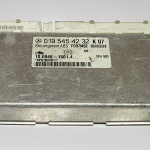 Op de afbeelding: Een zilverkleurige elektronische regeleenheid met een wit label met tekst en barcodes. De eenheid heeft een rechthoekige vorm met een metalen behuizing en twee witte connectoren. Het label bevat de tekst "Steuergeraet ABS FD97M02".