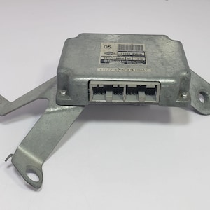 May include: A silver-colored automotive electronic control unit (ECU) with a bracket. The ECU has a rectangular shape with multiple connectors and a label with text and a barcode. The bracket has multiple holes for mounting.