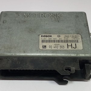 Puede incluir: Una unidad de control electrónico Motronic gris con una etiqueta Bosch y el texto "Made in Germany". La unidad tiene forma rectangular con soportes de montaje y un conector.