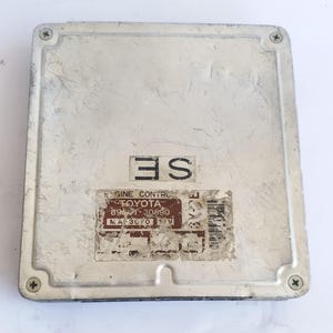 Puede incluir: Módulo de control del motor plateado, de forma rectangular. Tiene una etiqueta con el texto "ENGINE CONTROL TOYOTA" y otros números. Cuatro tornillos aseguran la cubierta. El módulo tiene un aspecto desgastado.