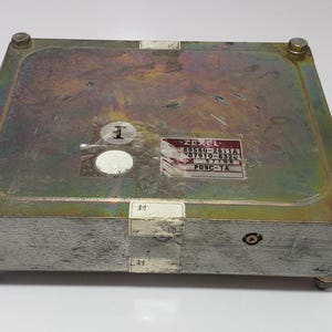 以下が含まれることがあります： 金属製の長方形の電子部品。表面はわずかに劣化しています。本体はシルバーで、上部はゴールド色で、ボルトで固定されています。テキストが書かれたステッカーがいくつか貼られています。おそらく自動車部品です。