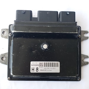 May include: A black Nissan engine control unit (ECU) with three black connectors. The unit has a silver metal frame with mounting tabs. A white label displays the text "NISSAN", "H 8", and a barcode. The text "Hitachi, Ltd. Tokyo Japan" is also visible.