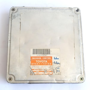 May include: A silver Toyota Emission Control module, rectangular in shape. It features a sticker with the Toyota logo and other text, including a barcode. The module is secured by four screws at the corners.