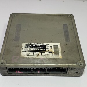 May include: A gray Toyota engine control module with a rectangular shape and multiple connection ports. The module has a sticker with text including "ENGINE CONTROL TOYOTA" and model numbers. The module is designed for a 12V system.