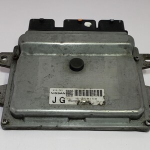 May include: A silver Nissan engine control unit (ECU) with black connectors and a black button. The unit is rectangular with mounting tabs and a label with text and a barcode. The label reads "Nissan" and "Hitachi".