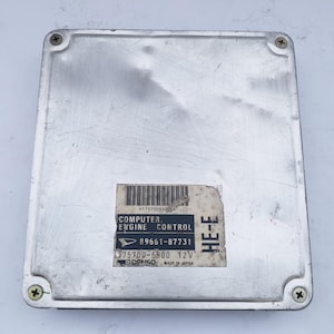 Puede incluir: Módulo de control del motor de computadora de color plateado con forma rectangular. El módulo tiene una etiqueta con el texto "COMPUTER ENGINE CONTROL" y "DENSO MADE IN JAPAN". Cuatro tornillos aseguran las esquinas.