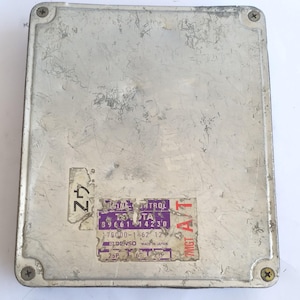 May include: A rectangular, silver-colored automotive computer module with visible wear and tear. It features a sticker with Japanese text and the brand name "DENSO." The module is secured with four screws at each corner.