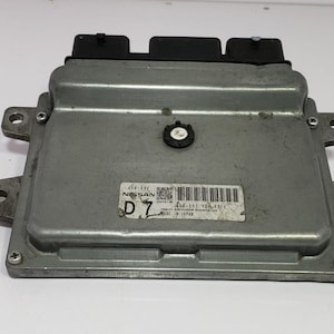 May include: A silver Nissan engine control unit (ECU) with black connectors. The ECU is rectangular with mounting tabs and a label that reads "NISSAN" and "MADE IN JAPAN".