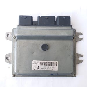 May include: A silver automotive computer module with three black connectors. The module has a label with the brand name "NISSAN" and a barcode. The text "9 A" is visible on the label. It has mounting tabs. The module is likely for a car.