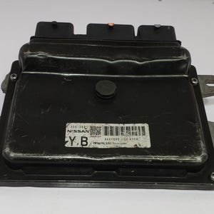 May include: A black electronic control unit (ECU) with a rectangular shape. The ECU has three black connectors on top and a label with text and a barcode. The unit has mounting brackets on the sides, likely for a vehicle.