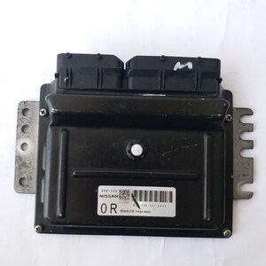 May include: A black Nissan engine control module with two black connectors. The module has a silver mounting bracket and a label with text and a barcode. The label reads "NISSAN" and "Hitachi Ltd Tokyo Japan".