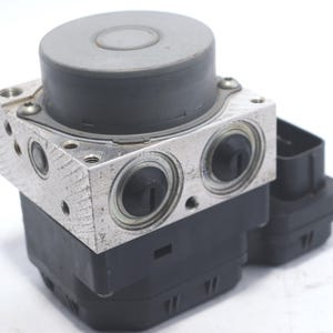 Op de afbeelding: Een zilver en zwart auto-onderdeel met een cilindrische bovenkant en meerdere poorten. Het onderdeel heeft een rechthoekige basis en een zwarte connector aan de zijkant. De component lijkt gemaakt van metaal en plastic.