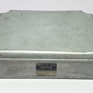 以下が含まれることがあります： シルバーカラーのトヨタコンピューターアセンブリバッテリー。長方形の金属製の箱は角が丸く、ネジで固定されています。前面にはテキストのラベルが見えます。白い表面に置かれています。