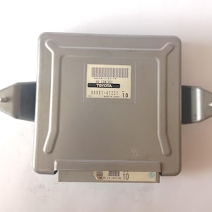 May include: A silver-colored Toyota HV Control module with a rectangular shape. The module has a label with the text "HV CONTROL TOYOTA" and the part number "89881-47221". The module has mounting brackets on each side.