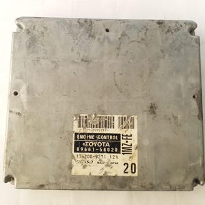 May include: A rectangular, silver engine control unit with a label that reads "ENGINE CONTROL TOYOTA" and "1MZ-FE". The unit has several mounting holes and the number "20" printed on the side.