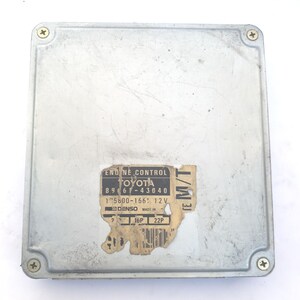 May include: A silver engine control module with four corner screws. The module has a label with the text "ENGINE CONTROL TOYOTA" and other technical details. This module is likely for a motor vehicle.