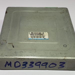 以下が含まれることがあります： 「MD339903  E2T45073 M 0029  MITSUBISHI ELECTRIC CORP. JAPAN」と記載されたラベルが付いたグレーの金属製自動車制御ユニット。ユニットには「MD339903」という青い文字が刻まれています。