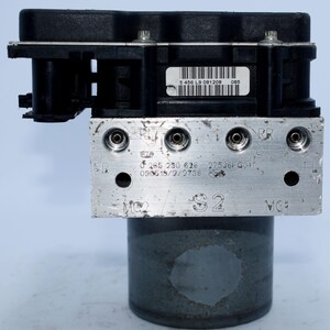 Op de afbeelding: Een ABS-module (Anti-blokkeersysteem) met een zwart kunststof bovendeel en een zilverkleurige metalen basis. De module heeft meerdere poorten en is gemarkeerd met verschillende codes en nummers. De cilindrische basis is donkergrijs.
