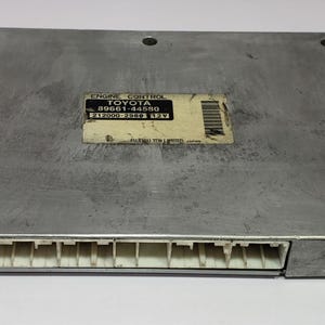May include: A silver Engine Control Unit (ECU) for a Toyota vehicle. The rectangular unit features a row of white connectors along one edge. A label with the Toyota logo and part number is affixed to the top.
