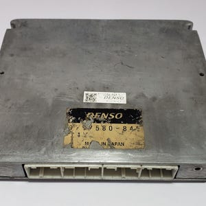 以下が含まれることがあります： 「DENSO 580-84」と記載された黒と黄色のラベルが付いた銀色の自動車制御ユニット。ラベルには「MADE IN JAPAN」の文字も記載されています。