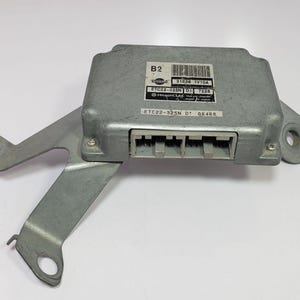 May include: A silver-coloured automotive electronic control unit (ECU) with a metal bracket. The ECU has multiple connectors and labels with text and a barcode. The bracket has mounting holes. The unit is likely for a Nissan vehicle.