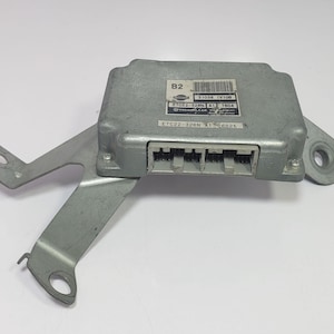 May include: A silver-colored automotive electronic control unit (ECU) with a metal bracket attached. The ECU has a rectangular shape with a series of connectors and a label with text and a barcode. The bracket has multiple mounting holes.
