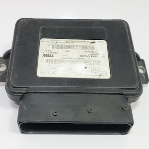 Puede incluir: Una unidad de control electrónico (ECU) negra con forma rectangular y un conector grande. La ECU tiene una etiqueta con texto y códigos de barras. La unidad es probablemente para aplicaciones automotrices o industriales.