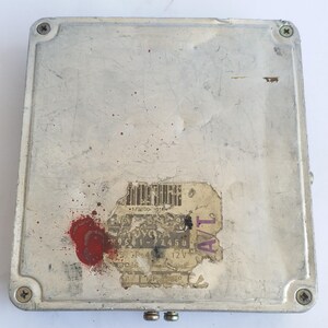 May include: A rectangular, silver automotive computer module with a worn appearance. The module has a sticker with text and a red stain. Four screws secure the top. Two small metal connectors are visible at the bottom.