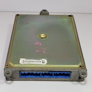 Könnte beinhalten: Ein rechteckiges elektronisches Steuergerät (ECU) mit goldfarbener Oberseite und blauem Stecker. Das Steuergerät hat ein Metallgehäuse mit Schrauben an jeder Ecke und ein kleines Etikett mit Barcode. Rosa handschriftlicher Text ist oben sichtbar.