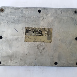 May include: A rectangular, silver engine control module with a sticker displaying "ENGINE CONTROL TOYOTA 89666-28140 211000-8940 12V". The module has several screw holes and is likely a car part.