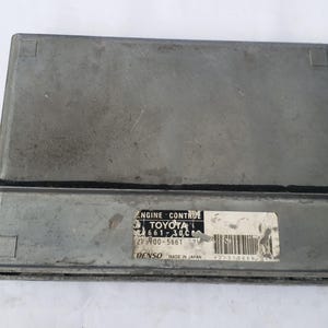 May include: A silver-colored Engine Control Unit (ECU) for a Toyota vehicle. The ECU has a rectangular shape with a black rubber seal and a label that reads "ENGINE CONTROL TOYOTA". The label also includes the text "MADE IN JAPAN".