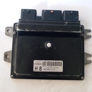 May include: A black Nissan engine control unit (ECU) with three black connectors. The ECU is rectangular with mounting tabs and a label reading "NISSAN H8". The unit is secured with screws.