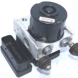 Op de afbeelding: Een auto-ABS-module, met een zwart cilindrisch onderdeel op een zilverkleurige metalen basis. Een zwarte rechthoekige connector is aan de zijkant bevestigd. De module heeft diverse zilverkleurige fittingen en een barcode sticker.