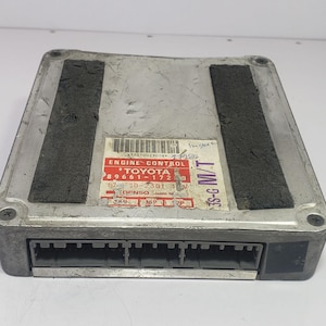 Könnte beinhalten: Ein silbernes Toyota Motorsteuergerät mit einem schwarzen Stecker und zwei schwarzen Schaumstoffpads. Das Modul hat ein rot-weißes Etikett mit dem Text "ENGINE CONTROL TOYOTA".