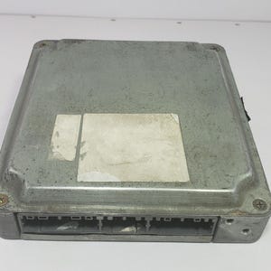 以下が含まれることがあります： 長方形の銀色の電子制御ユニット（ECU）で、片側にコネクタが付いています。 ECUはわずかにテクスチャ加工された表面と色あせた白いステッカーが付いています。留め具がユニットの角を固定しています。