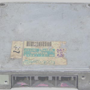 May include: A silver Engine Control Unit (ECU) with a rectangular shape. The ECU features a label with the text "ENGINE CONTROL TOYOTA" and a barcode. The unit has multiple connector ports and is secured with screws.