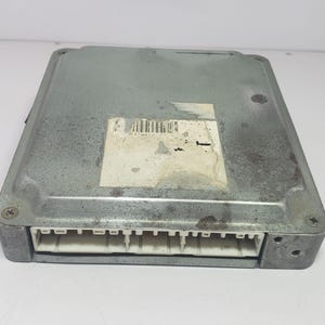 May include: A silver-coloured electronic control unit (ECU) with a rectangular shape. The ECU has a white connector on one side and a sticker with a barcode. The metal casing shows signs of wear and tear.