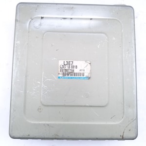 Puede incluir: Una unidad de control electrónico (ECU) rectangular de metal gris claro. Una etiqueta blanca con texto negro está adherida en la parte superior, mostrando códigos y el logotipo de Mitsubishi Electric Corp. Japan. La ECU tiene una superficie ligeramente texturizada.