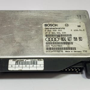 Puede incluir: Módulo de control Getriebesteuerung Bosch plateado con conector negro. El módulo tiene varias etiquetas con texto y códigos de barras. El texto incluye "BOSCH", "Made in Germany" y números de pieza. Módulo rectangular con pestañas de montaje.