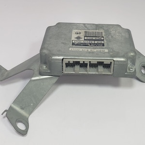 May include: A silver-colored automotive electronic control unit (ECU) with a mounting bracket. The ECU has multiple connectors and a label with text and a barcode. The bracket has multiple mounting holes.