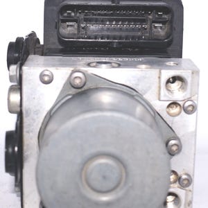 Op de afbeelding: Een zilverkleurig auto-onderdeel met een zwarte connector aan de bovenkant. Het onderdeel heeft een cilindrisch component en is vastgezet met meerdere schroeven. De achtergrond is effen wit.